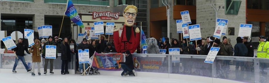 OPSEU members on strike, holding picket signs on ice rink next to Kathleen Wynne puppet