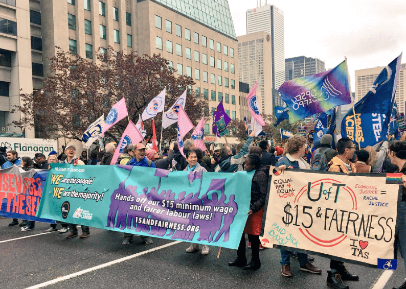 Crowd of people holding up flags and banners, including OPSEU flags, in support of $15 minimum wage and fair labour laws