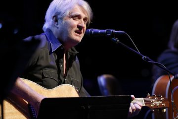 Tom Cochrane plays guitar and signs in to a microphone.