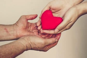 One person holding a heart in their hands, another set of hands underneath holding
