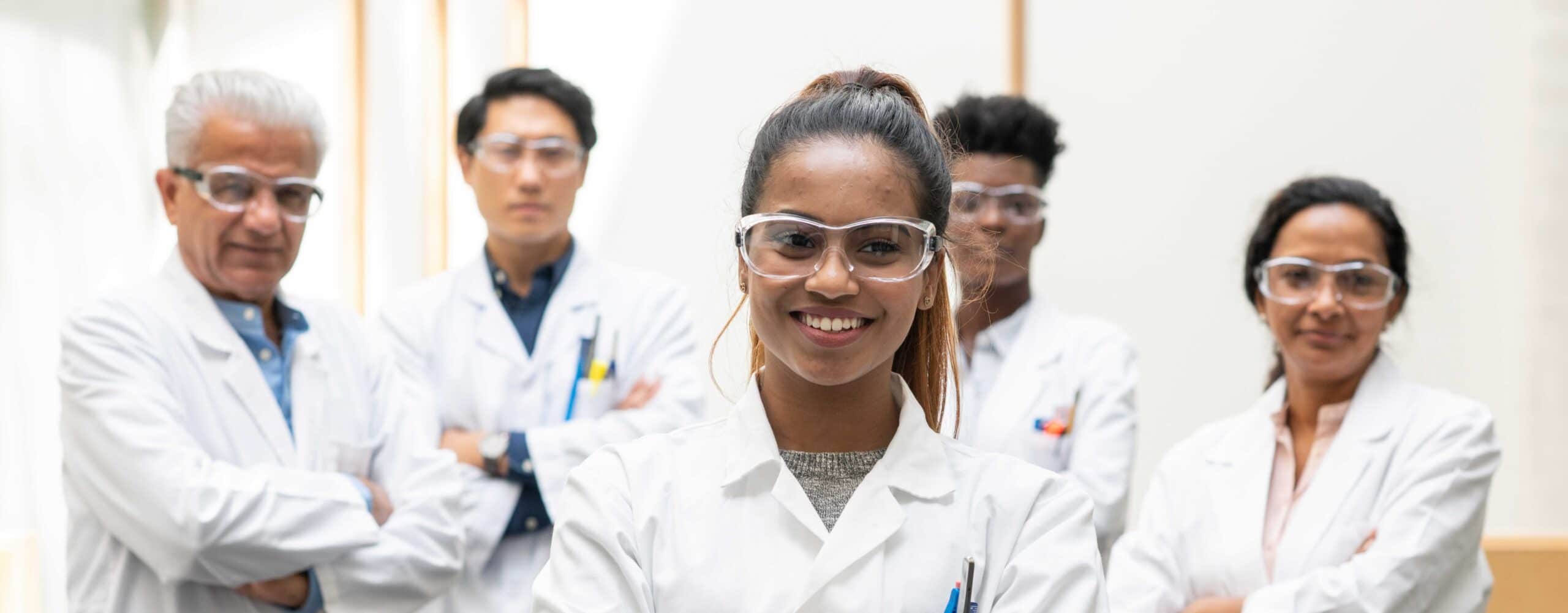 A multi-ethnic group of medical scientists stop and pause for a team portrait before continuing their work. They are each smiling while wearing lab coats and protective eye wear for safety.