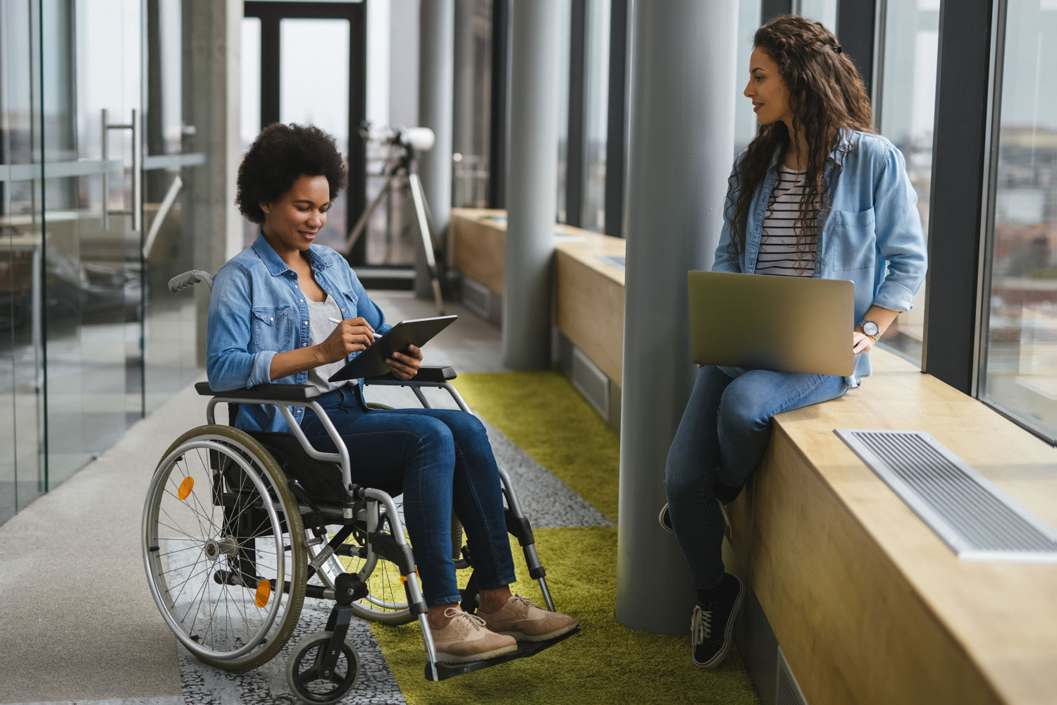 Woman in a wheelchair uses a tablet as another woman uses a laptop