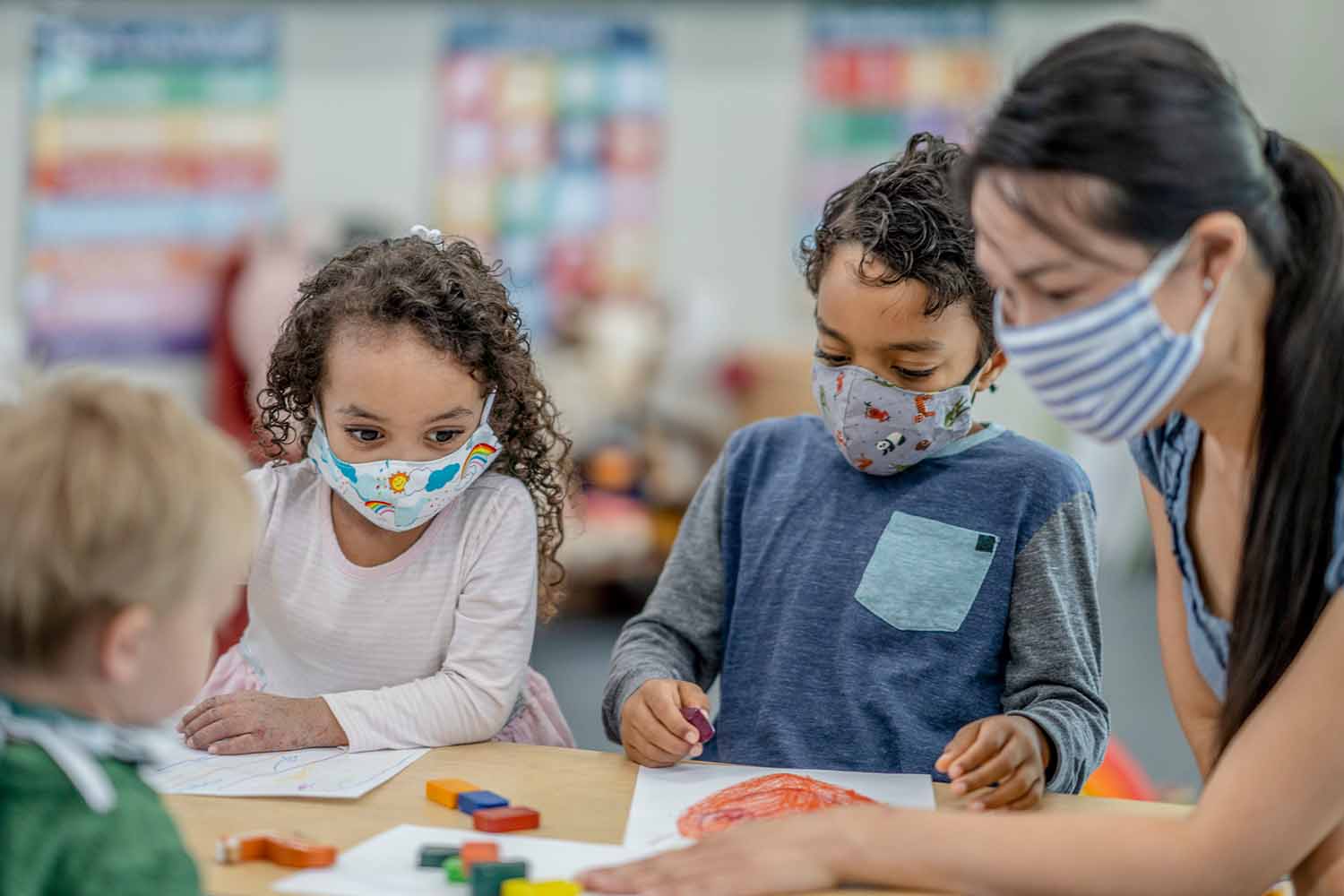 Multi-ethnic group of children and childcare worker colouring at a table while wearing protective face masks to avoid the transfer of germs.