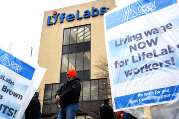 Striking LifeLabs workers in front of a building with a LifeLabs sign and a picket sign that reads "Living wage NOW for LifeLabs workers!"